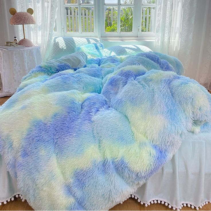 a bed covered in blue and green fluffy blankets next to a window with white curtains