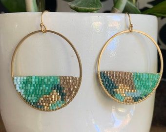 a pair of gold hoop earrings with blue and green seed beads on them sitting next to a potted plant