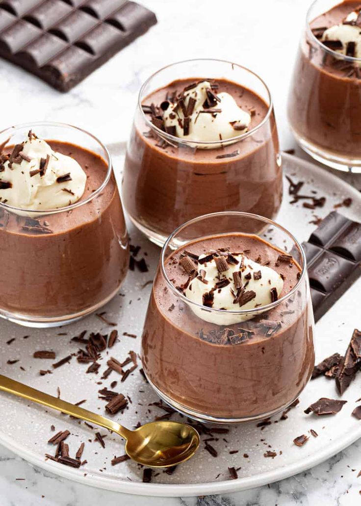 three glasses filled with chocolate pudding on top of a white plate next to two spoons