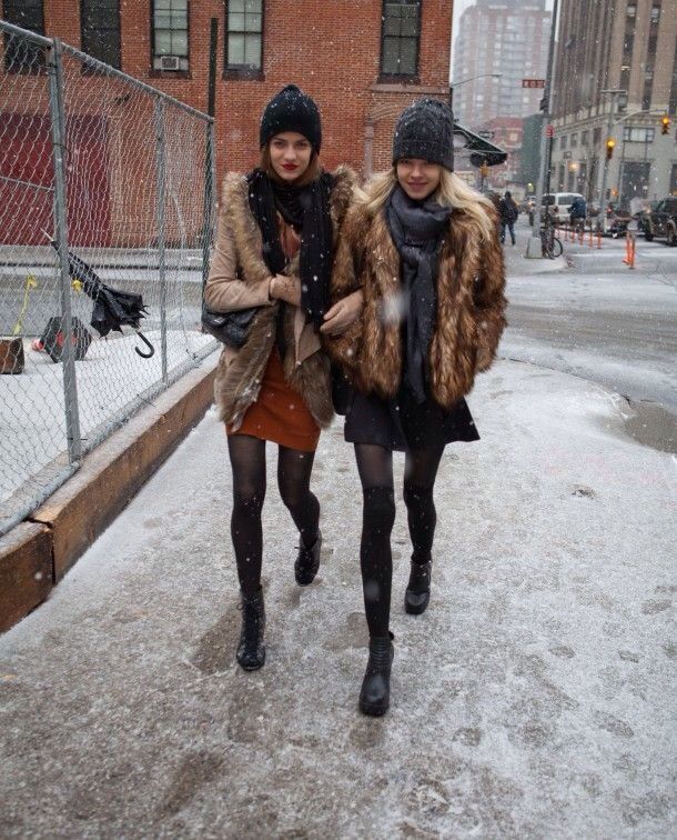 two women walking down the street in the snow wearing winter clothes and fur vests