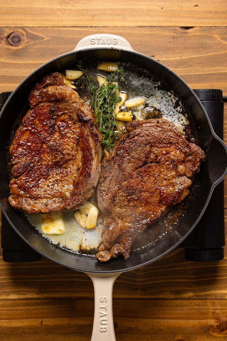 two steaks cooking in a skillet on top of a wooden table next to a knife and fork