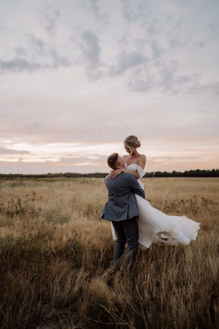 a bride and groom embracing in the middle of an open field at sunset with clouds overhead