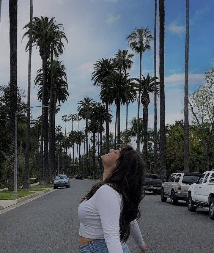 a woman standing in the middle of a street with palm trees and cars behind her