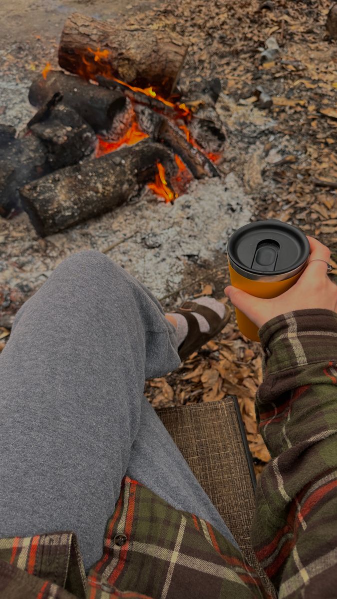 a person sitting in front of a campfire holding a cup with the lid up