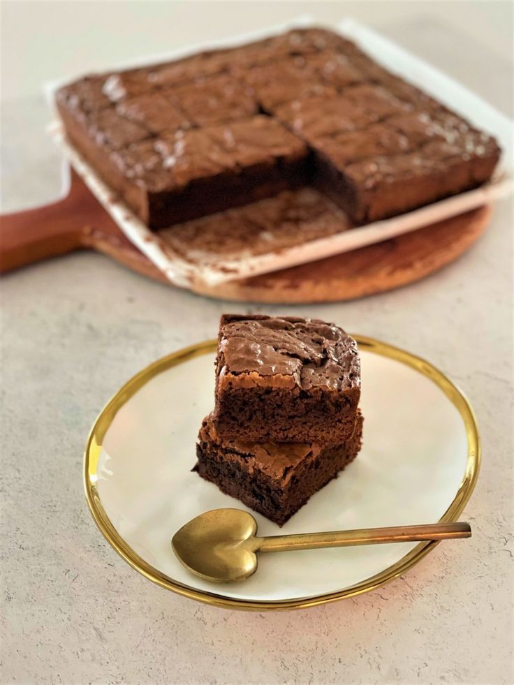 two pieces of brownie on a plate with a spoon next to it and another piece of cake in the background