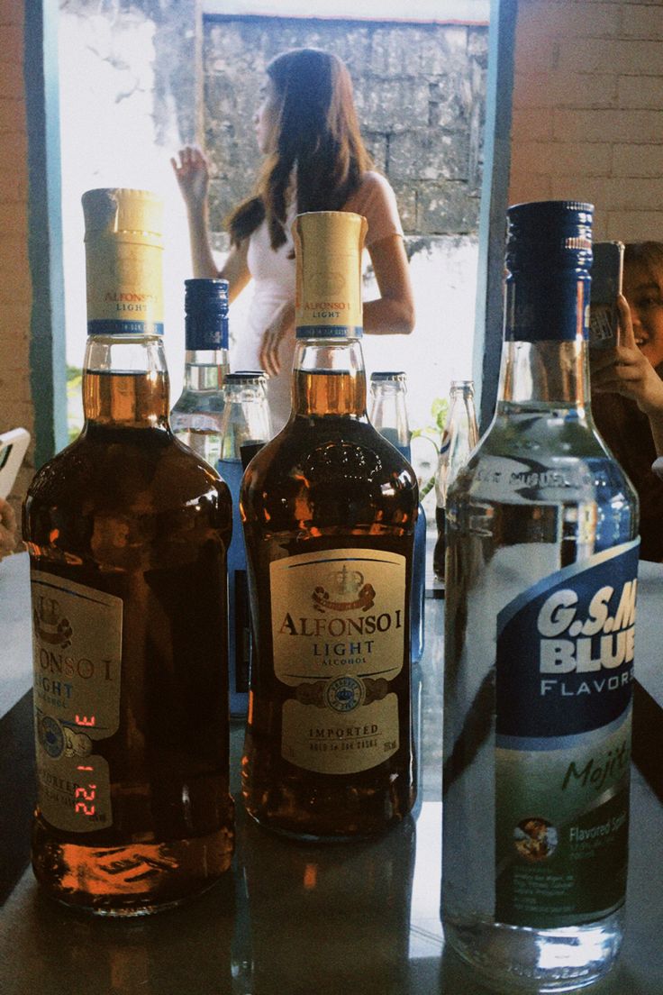 three bottles of alcohol sitting on top of a counter next to a woman looking out the window