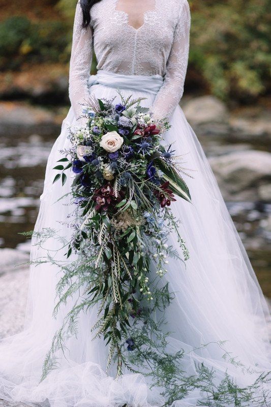 a woman in a wedding dress holding a bouquet with flowers on the side, standing next to a river