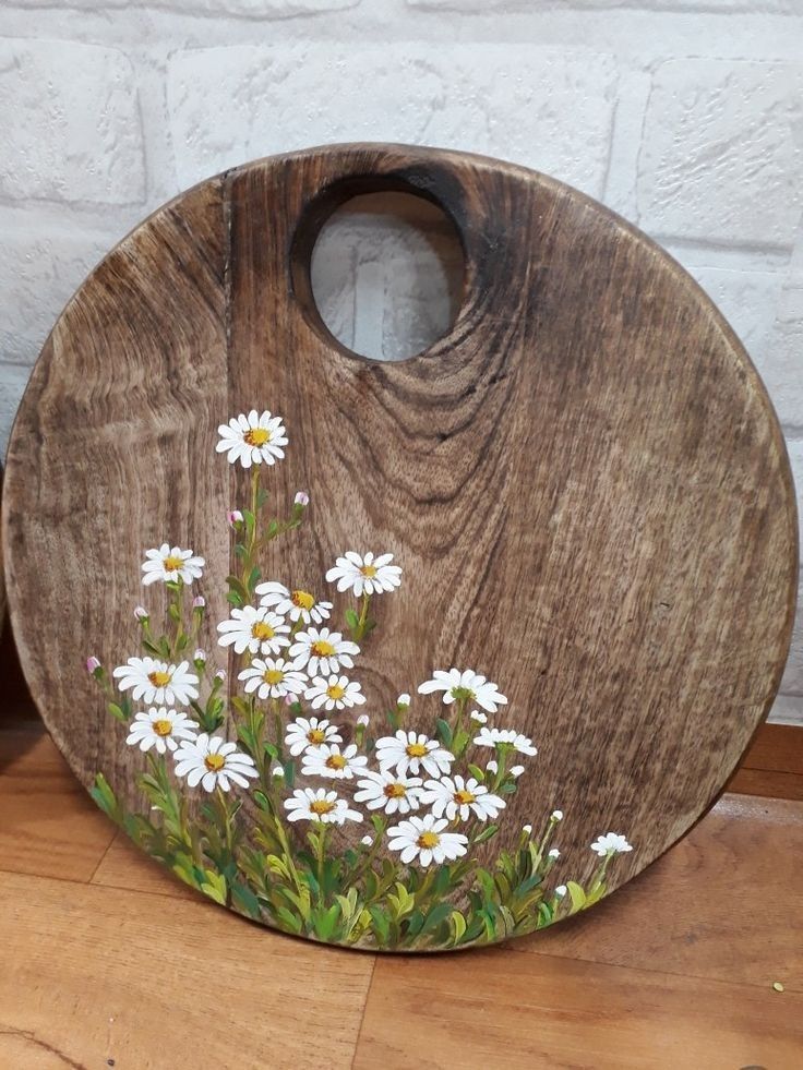 a wooden cutting board with white daisies painted on the front and bottom, against a brick wall