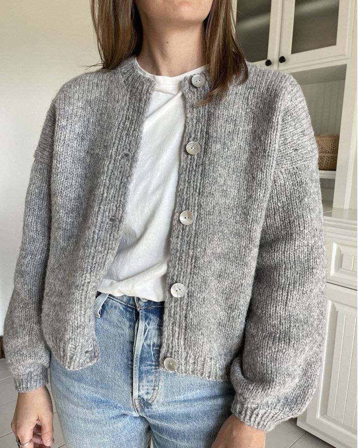 a woman standing in a kitchen wearing a gray cardigan sweater and blue denim jeans