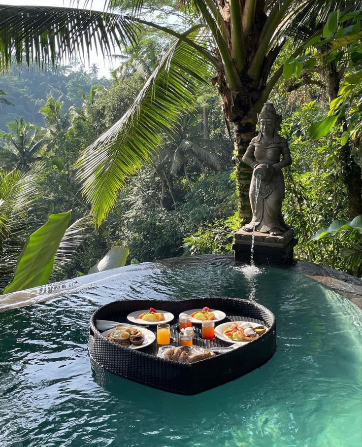 an outdoor pool with food on it and a statue in the background, surrounded by greenery