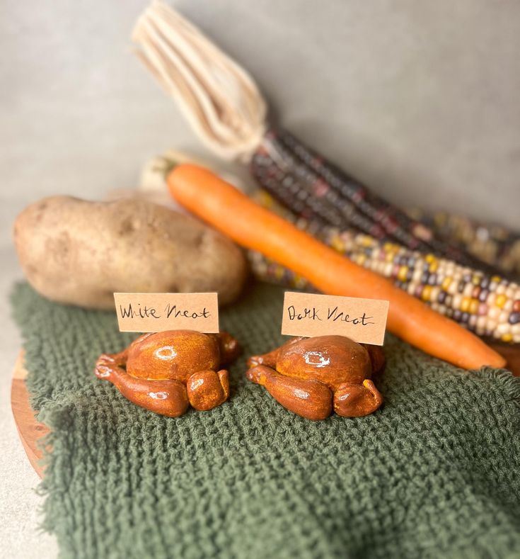 two little ceramic elephants sitting on top of a green cloth next to carrots and corn