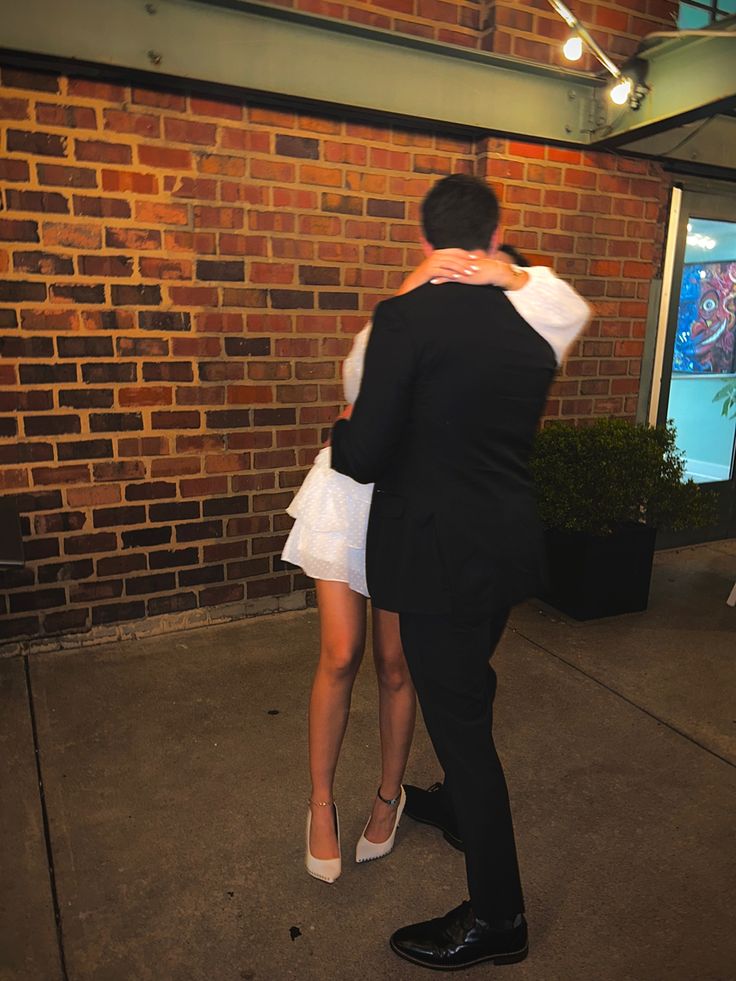 a man and woman in formal wear embracing each other on the sidewalk next to a brick wall