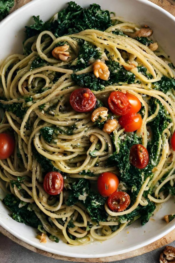 a white bowl filled with pasta and spinach covered in tomatoes, nuts and kale