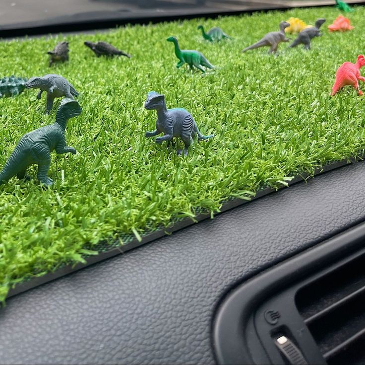 small toy dinosaurs in the grass on top of a car dashboard dash mat, with green grass behind them