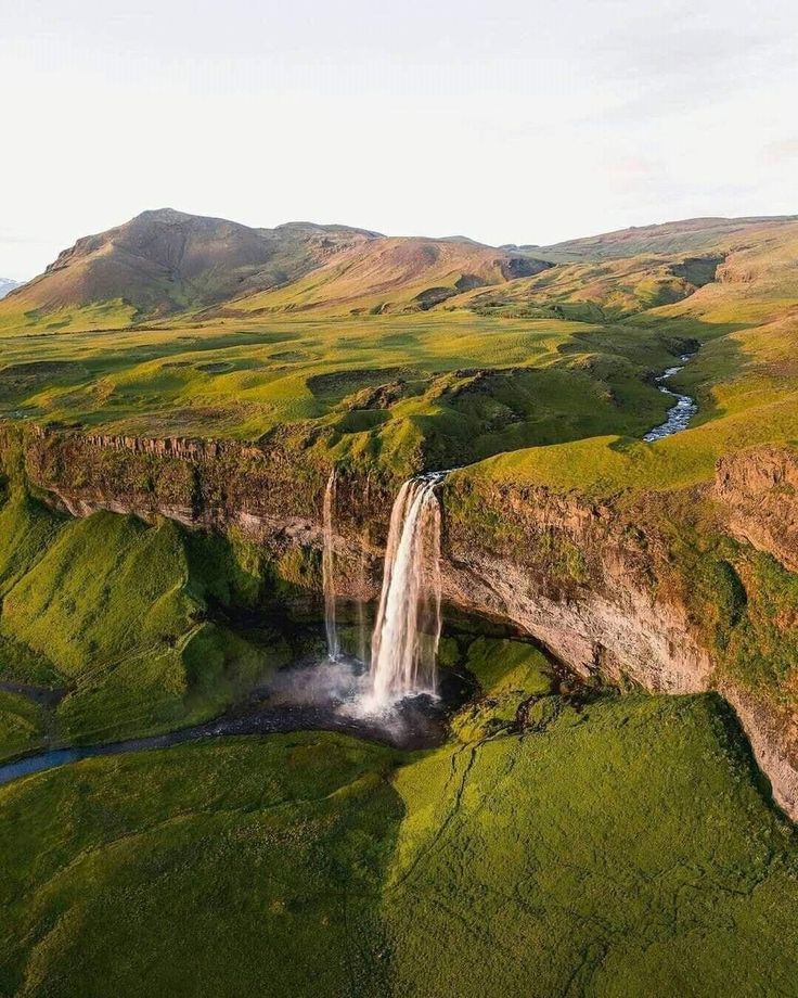 an aerial view of a waterfall in the middle of green fields and mountains with water running down it