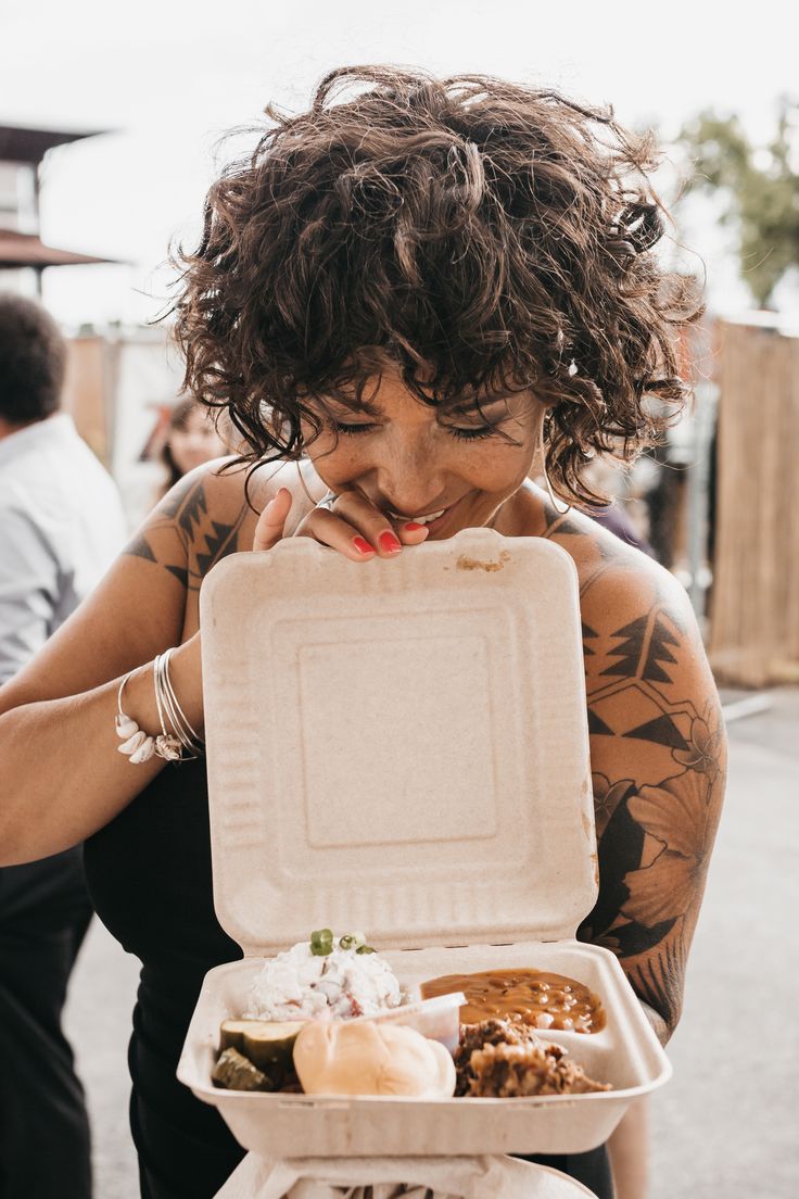 a woman holding a white box filled with food on top of her chest and looking at the camera