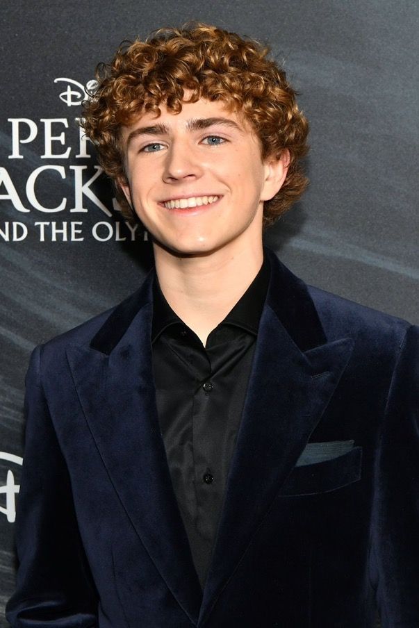 a young man with curly hair wearing a blue suit and black shirt smiling at the camera