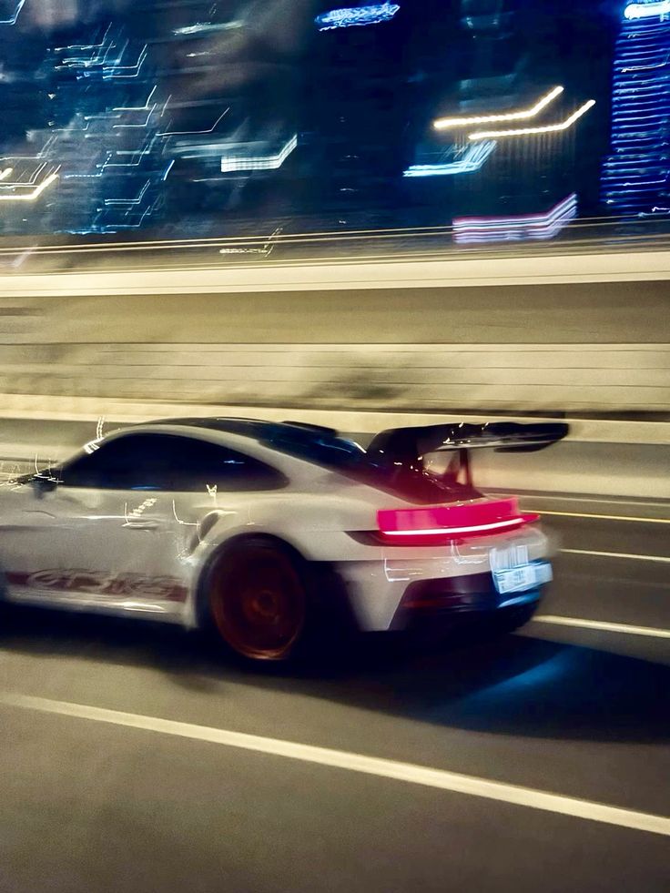 a white sports car driving down the road at night with city lights in the background