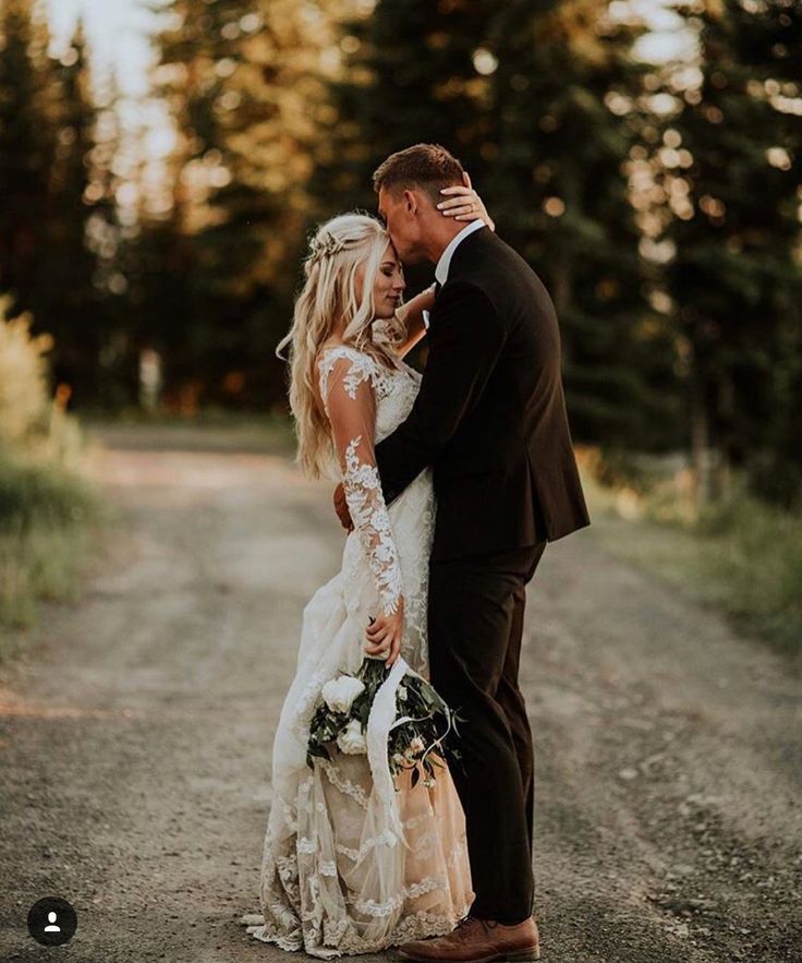 a bride and groom standing in the middle of a dirt road with trees behind them