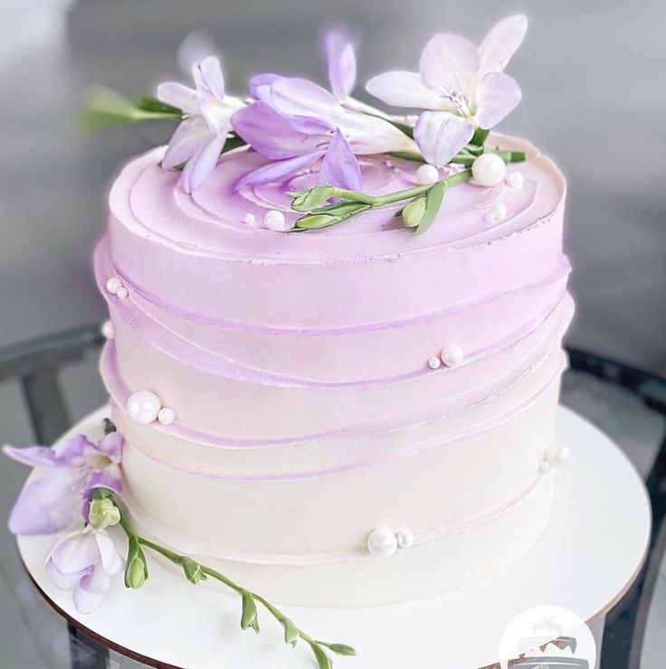 a white and purple cake with flowers on it sitting on a glass table in front of a window