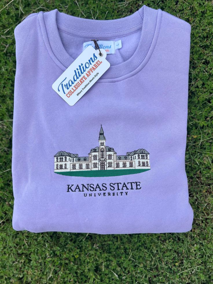 a purple kansas state university t - shirt laying on the grass with a tag hanging from it