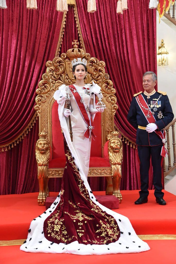 the queen is standing in front of an ornate throne with red drapes and gold trimmings