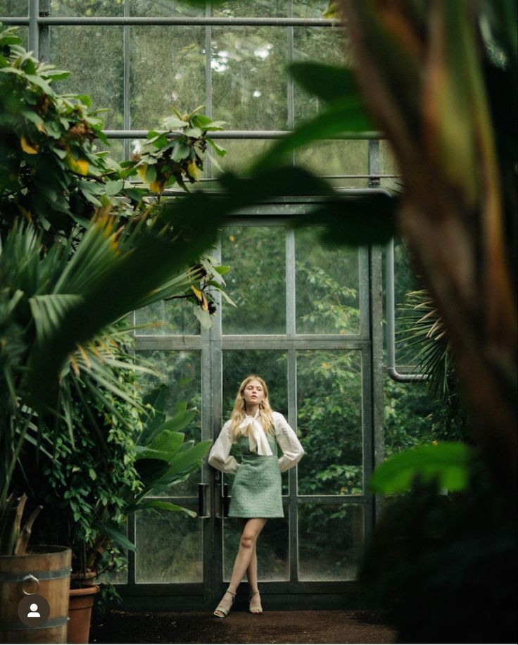 a woman standing in front of a window surrounded by plants and greenery with her hands on her hips