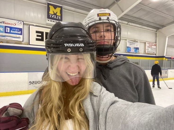 two people with helmets on taking a selfie in an ice rink while one person holds his hand out to the camera