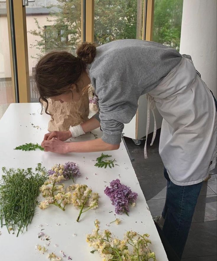 two people are arranging flowers on a table in front of a window, one woman is bending over and the other has her head down