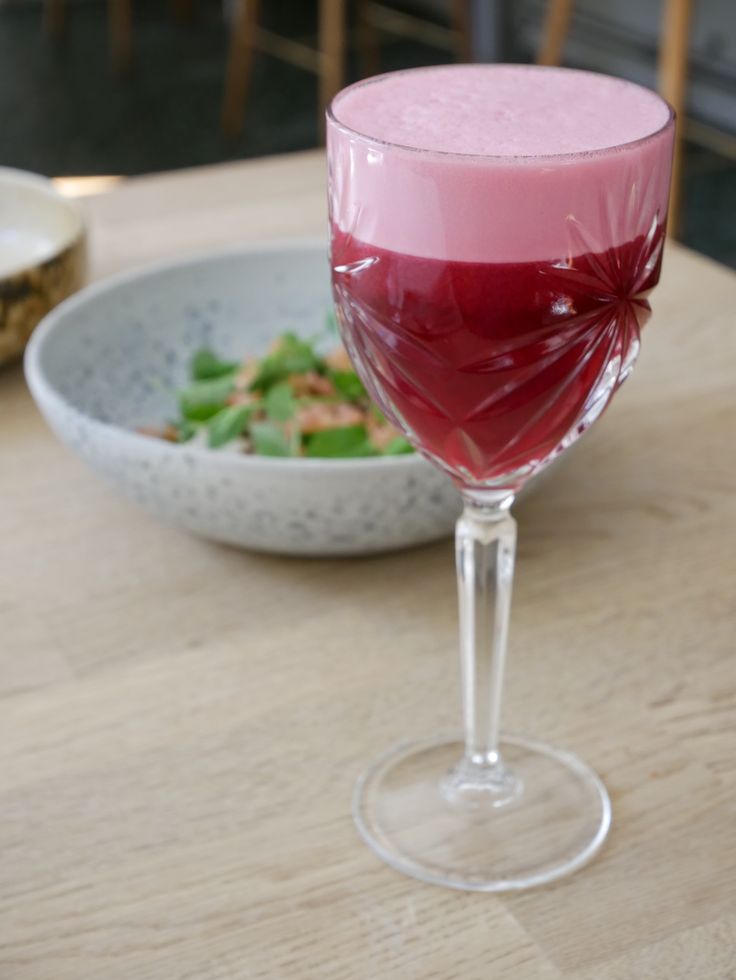 a pink drink in a wine glass next to a bowl of salad on a table