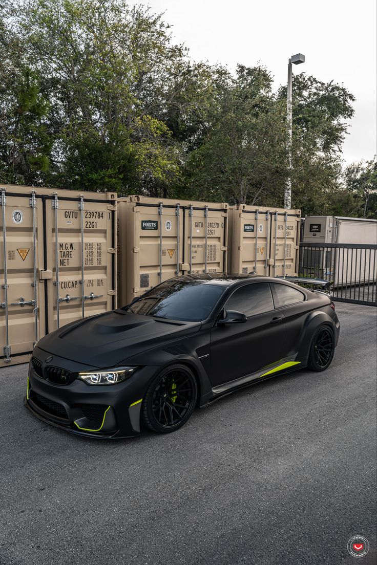 a black car is parked in front of some crates on the street with trees and bushes behind it