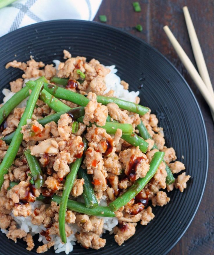 a black plate topped with meat and green beans next to chopsticks on a wooden table