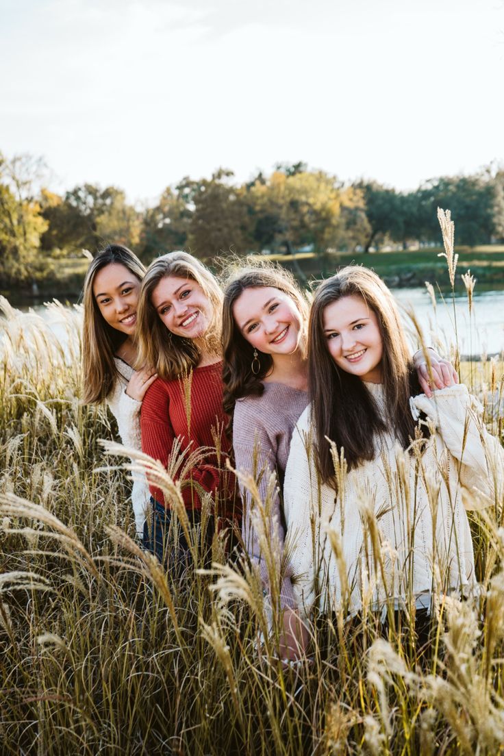 three girls are standing in tall grass by the water and smiling at the camera with their arms around each other