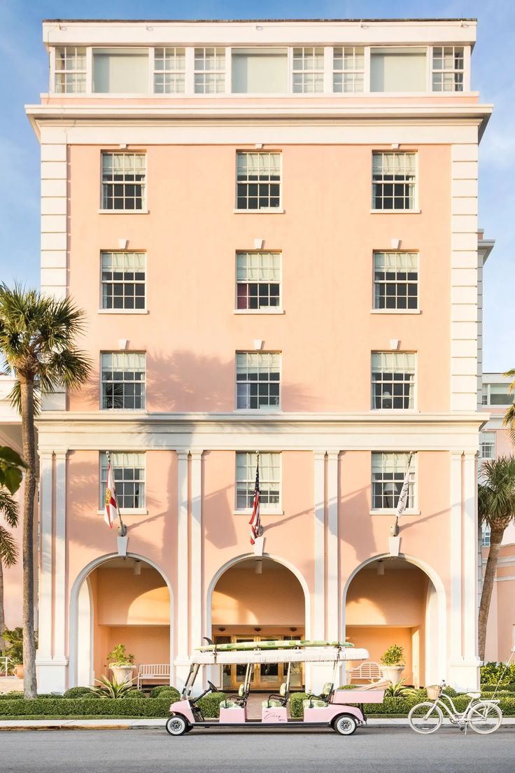 an old pink car is parked in front of a large building with palm trees on the side