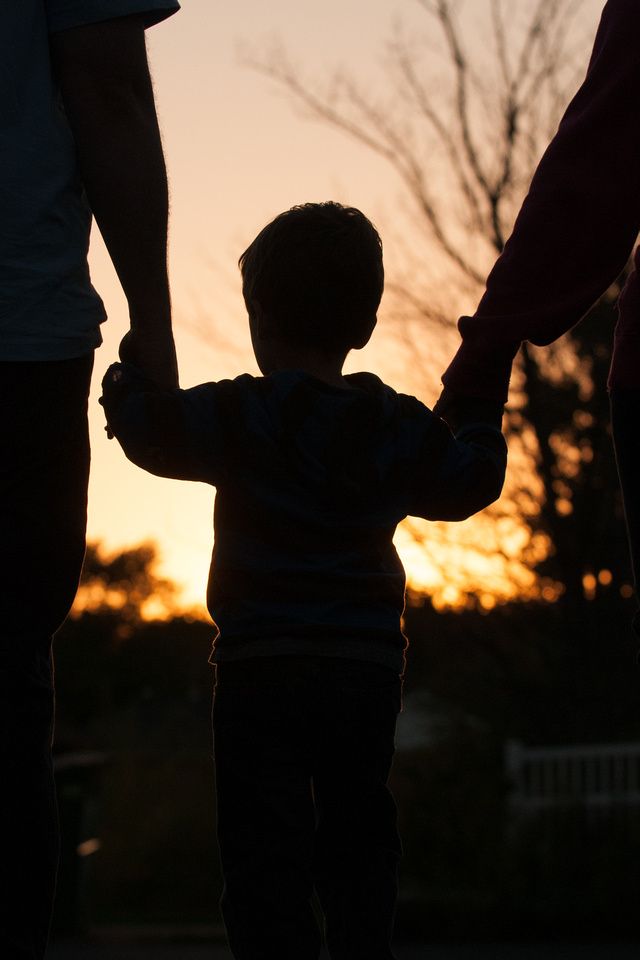 a little boy holding the hand of his mother and father as the sun sets in the background