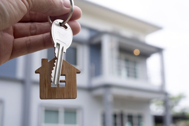 a hand holding a house key in front of a large white building with two story windows