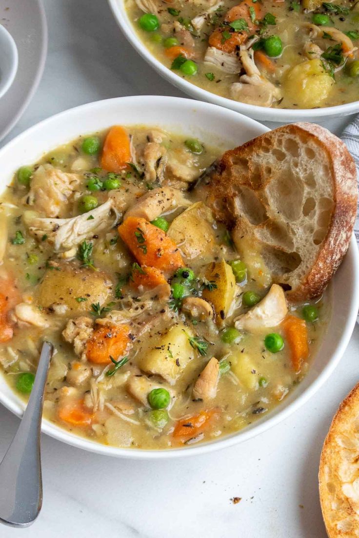 two white bowls filled with chicken and vegetable soup next to bread on a table top