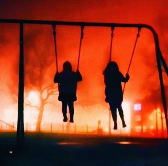 two people are sitting on swings in front of a fire that is burning behind them