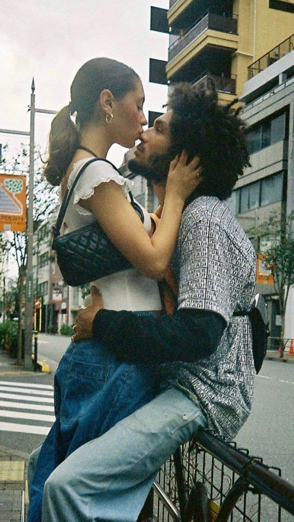 two people sitting on a fence kissing each other in the street with buildings behind them