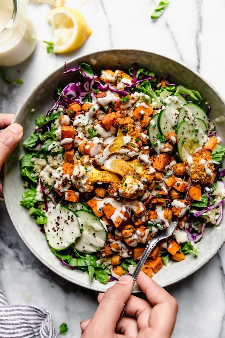 two hands holding a fork over a bowl of salad with dressing on it and cucumbers in the background