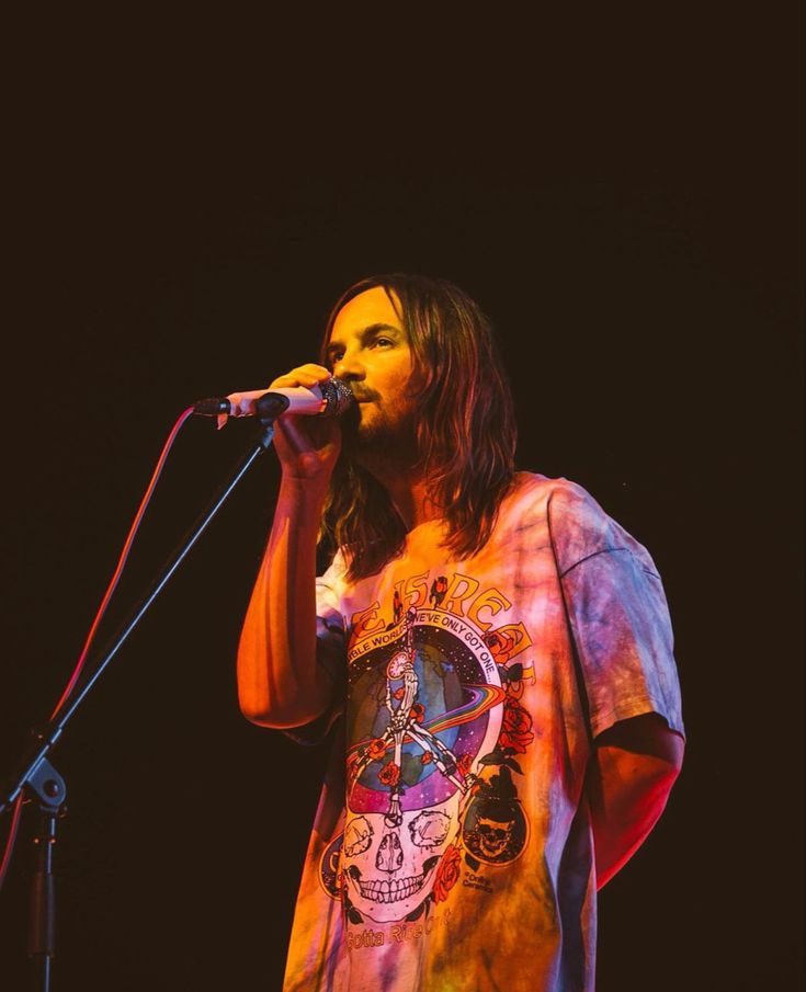 a man with long hair standing in front of a microphone and wearing a tie dye shirt