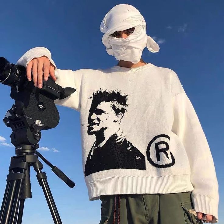 a man wearing a face mask and holding a camera next to a tripod in front of a blue sky