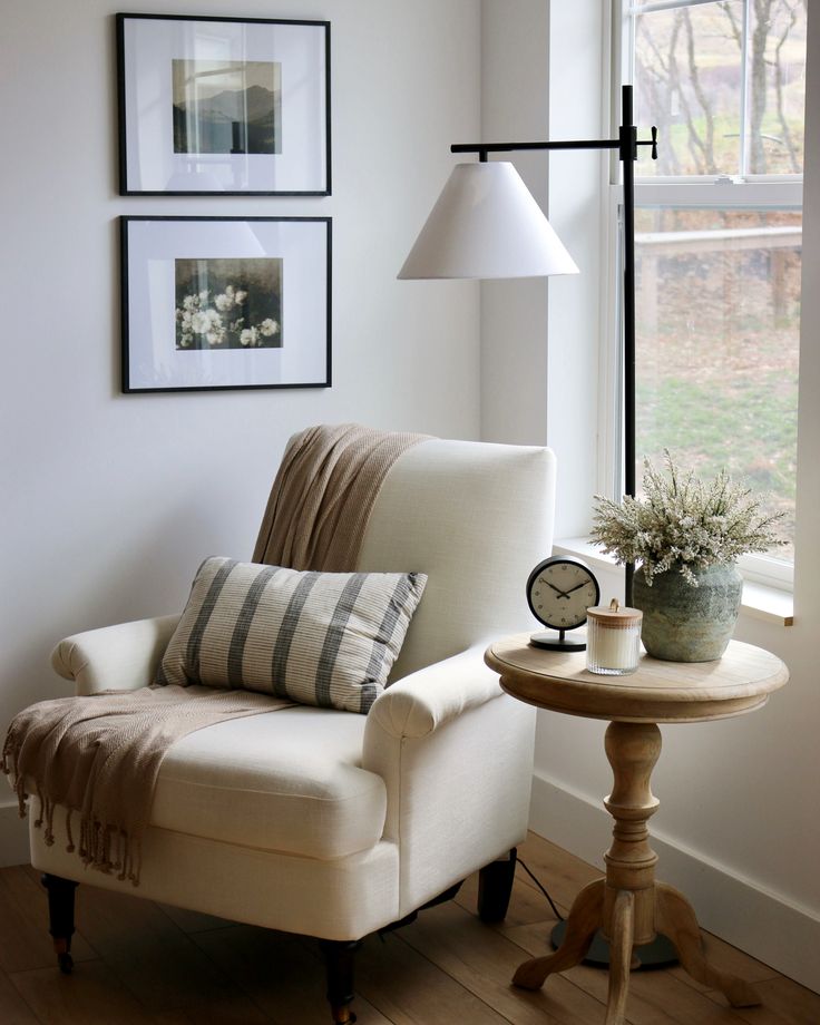 a white chair sitting next to a table with a lamp on it and pictures above