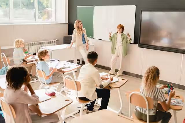 a group of children sitting at desks in front of a whiteboard with writing on it