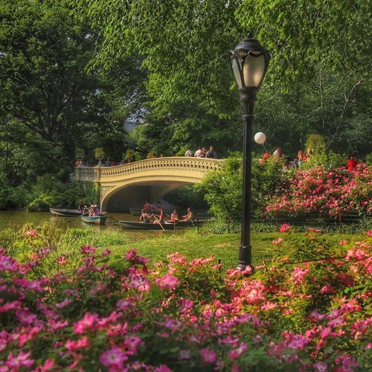 a lamp post in the middle of a park with flowers and people on boats going under a bridge