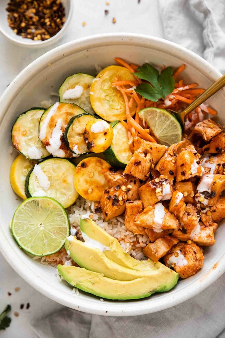 a white bowl filled with chicken, cucumber and rice topped with limes