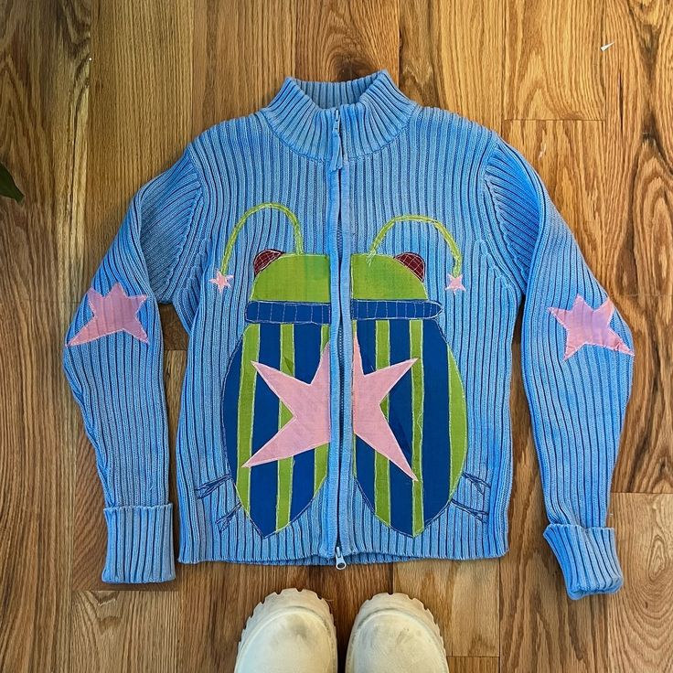 a child's blue sweater with pink stars and green beetle on the front, next to two pairs of white shoes