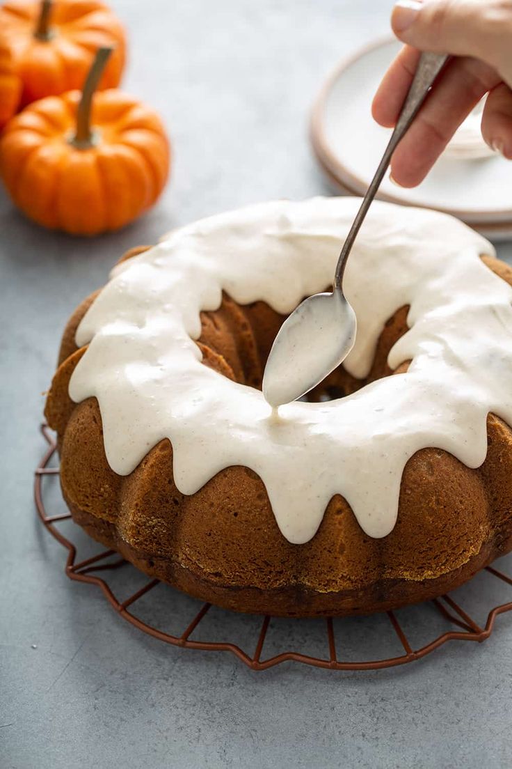 a person is spreading icing on a bundt cake with a fork and pumpkins in the background