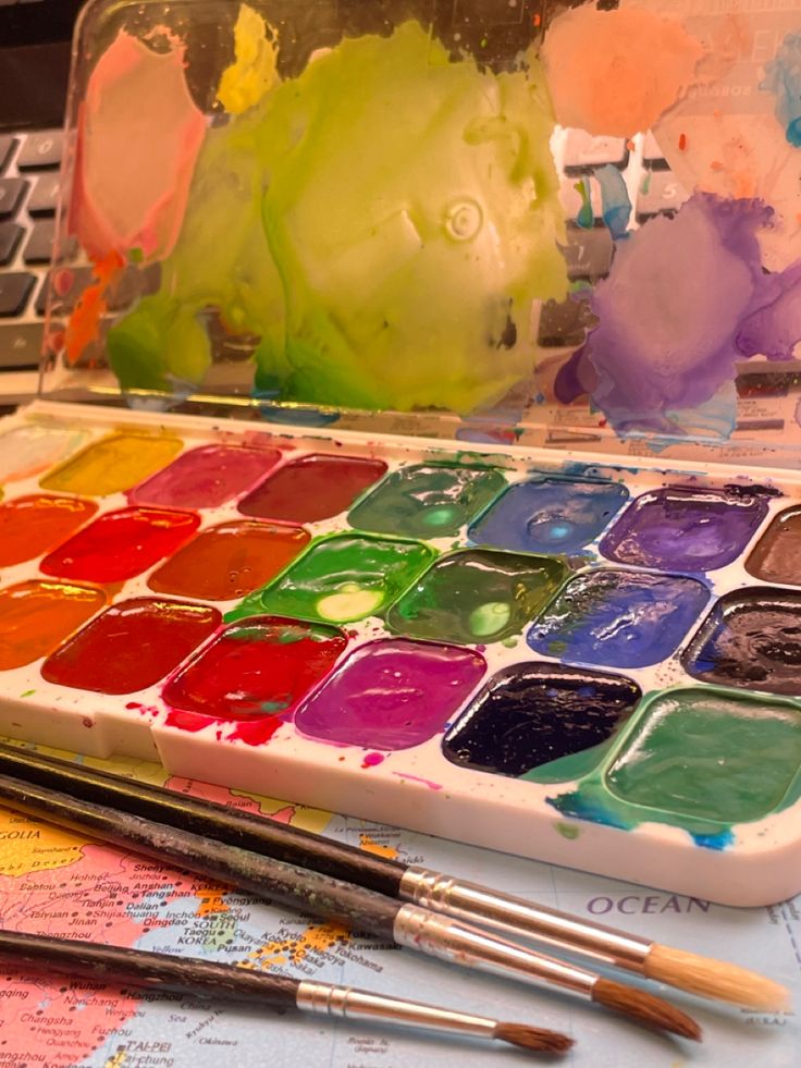 an open laptop computer sitting on top of a table covered in paint and watercolors