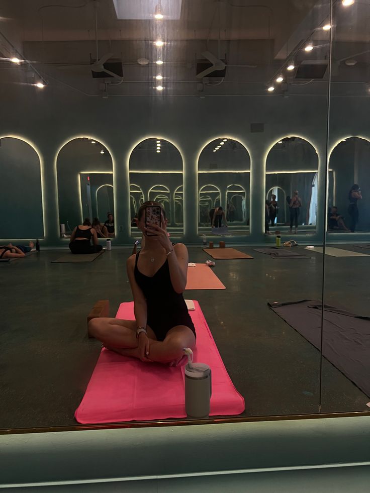 a woman sitting on a pink yoga mat in front of a glass wall and looking at her cell phone